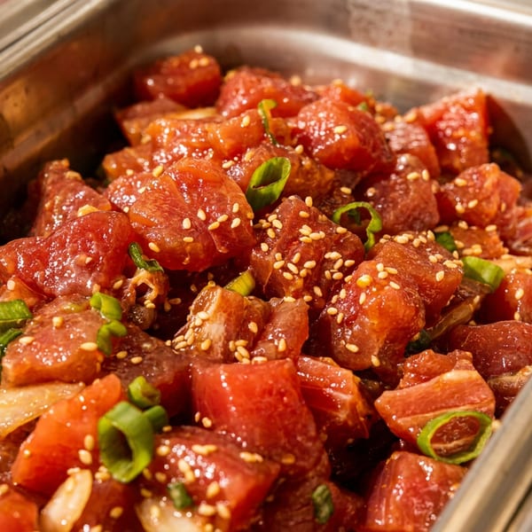 Close-up of a metal tray filled with glistening red poke, topped with sesame seeds and green onions.