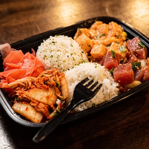 Japanese-inspired takeout bowl with seasoned rice, poke-style tuna, glazed chicken, kimchi, pickled ginger, and a black fork.