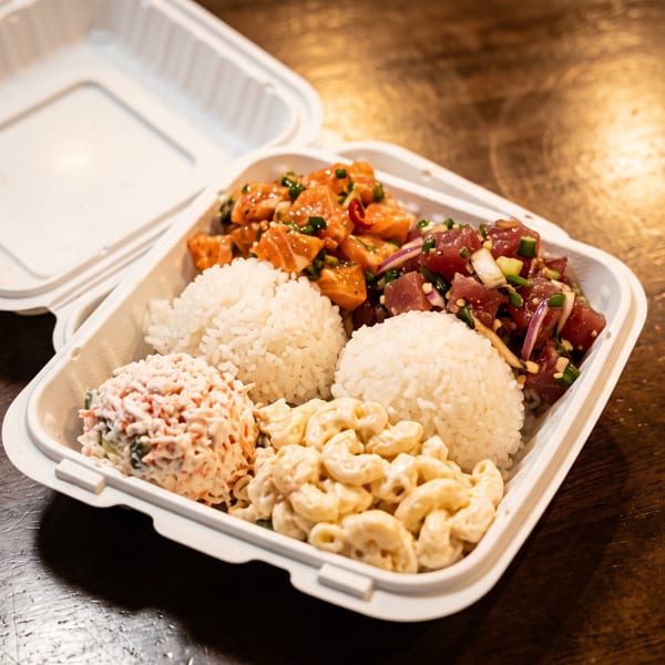 Takeout container filled with white rice, spicy salmon and tuna poke, macaroni salad, and creamy side dishes on a wooden table.
