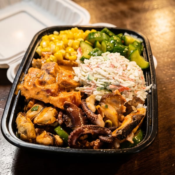 Takeout tray filled with sauced seafood, octopus, creamy slaw, corn, and cucumber salad on a wooden table.