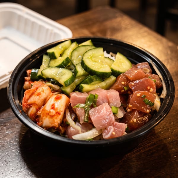 Black bowl of poke topped with sliced cucumbers, salmon, tuna, kimchi, onions, and scallions on a wooden table.